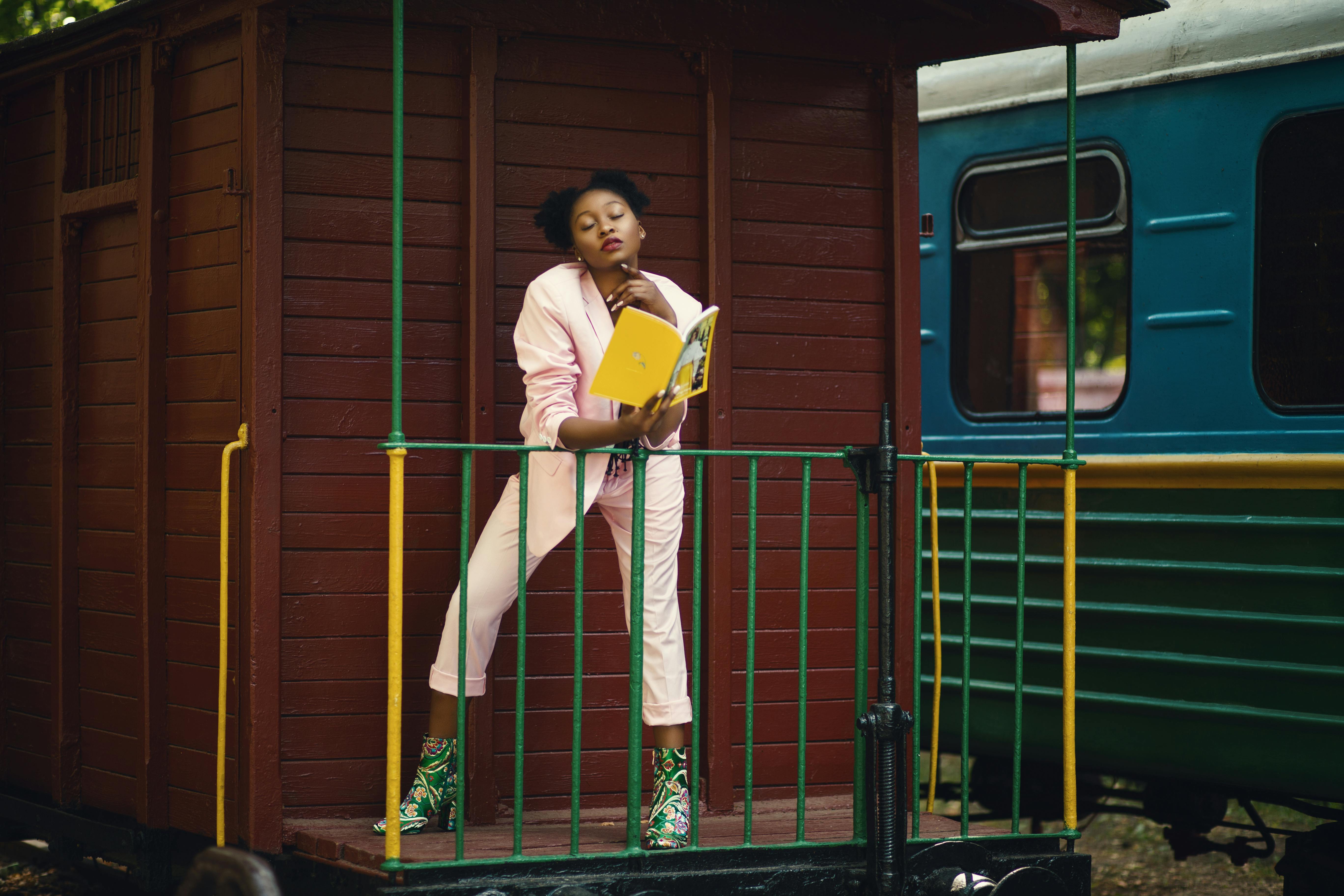 A young woman wearing pink blazer pants and green-heeled shoes holding a yellow-covered book