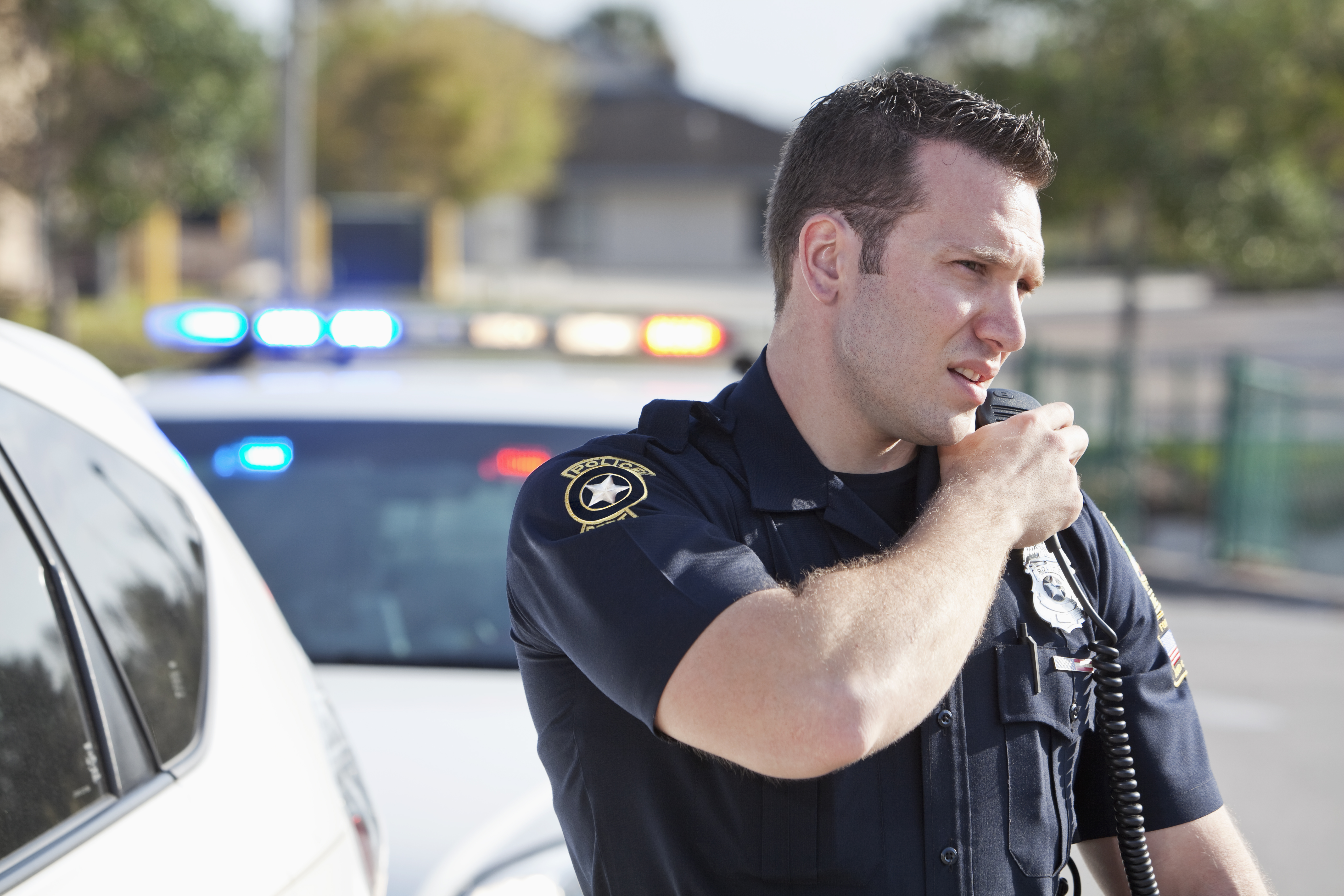 A male police officer standing outdoors, reaching for a radio.