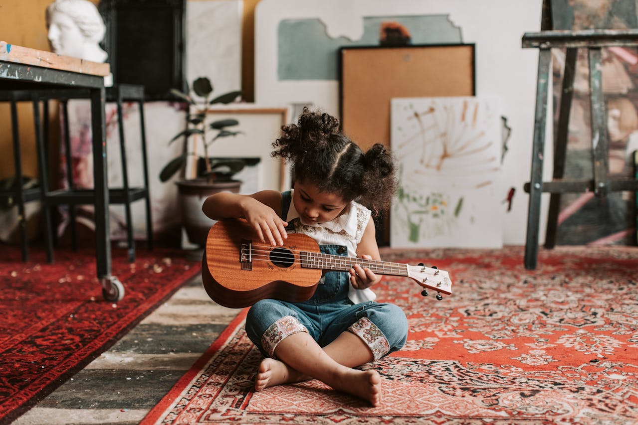 A young girl in a denim jumper sitting on the floor while playing a ukulele instrument