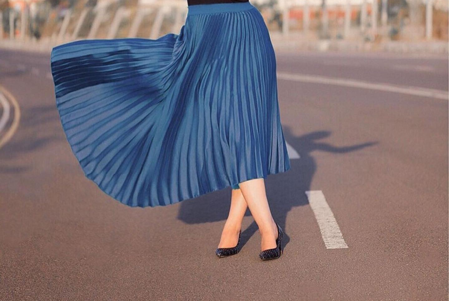 A woman stands on the road, wearing a blue pleated skirt
