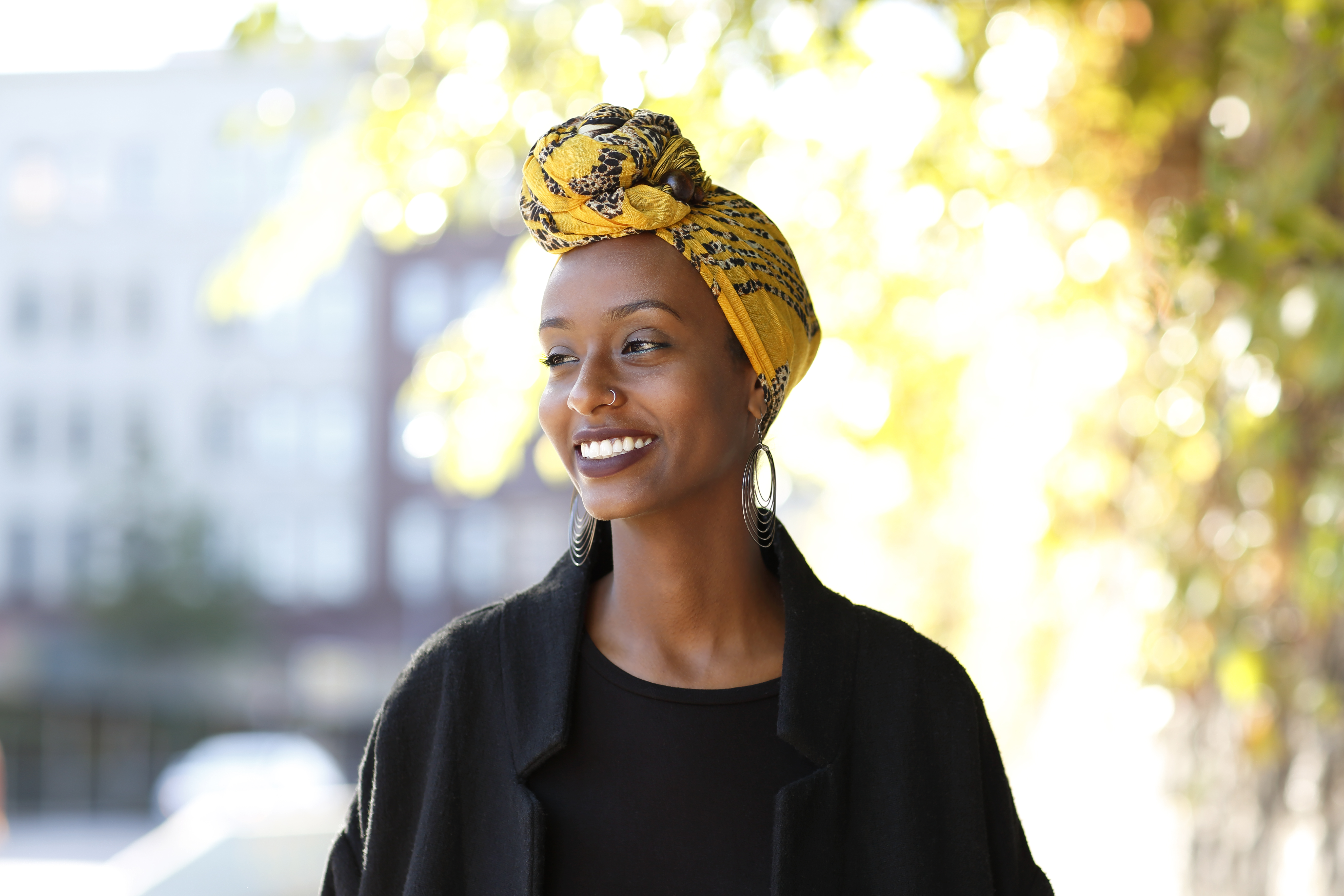 Beautiful, young, happy Muslim woman photographed in a bright outdoor urban setting wearing a yellow, decorated wrap around her head.