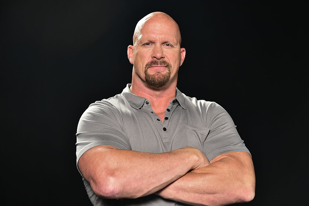 Steve Austin Stone Cold, one of the 90s wrestlers, is wearing a grey t-shirt and folding his hands as he poses for a photo.