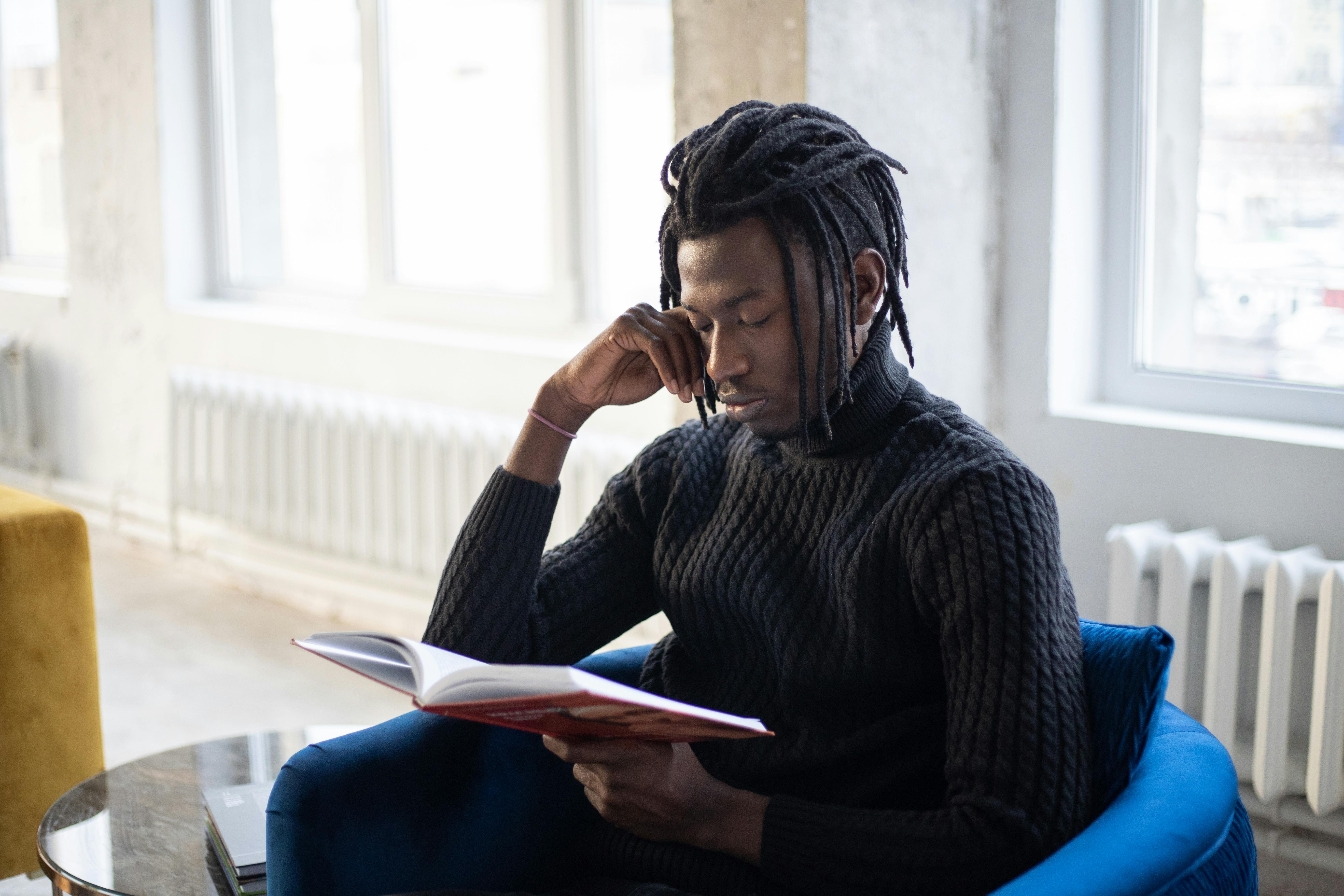 A young man in dreadlocks and a black sweater is reading a book