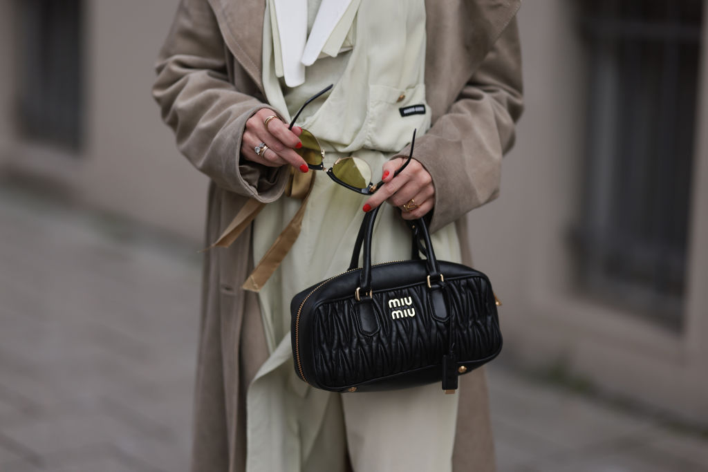 A stylish woman in a trench coat and sunglasses holding a black Miu Miu leather bag.