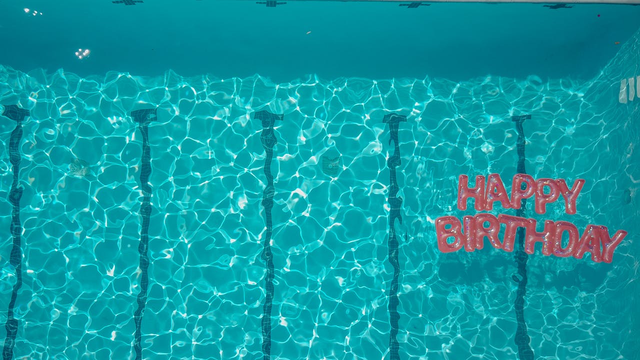 Bright "Happy Birthday" sign bobbing in a pool, with a backdrop of colourful pool floats.