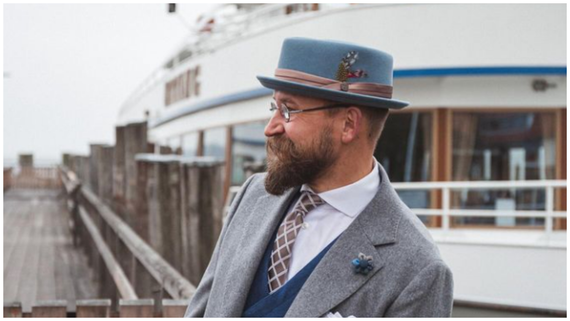 A man is standing next to yacht wearing a blue pork pie hat