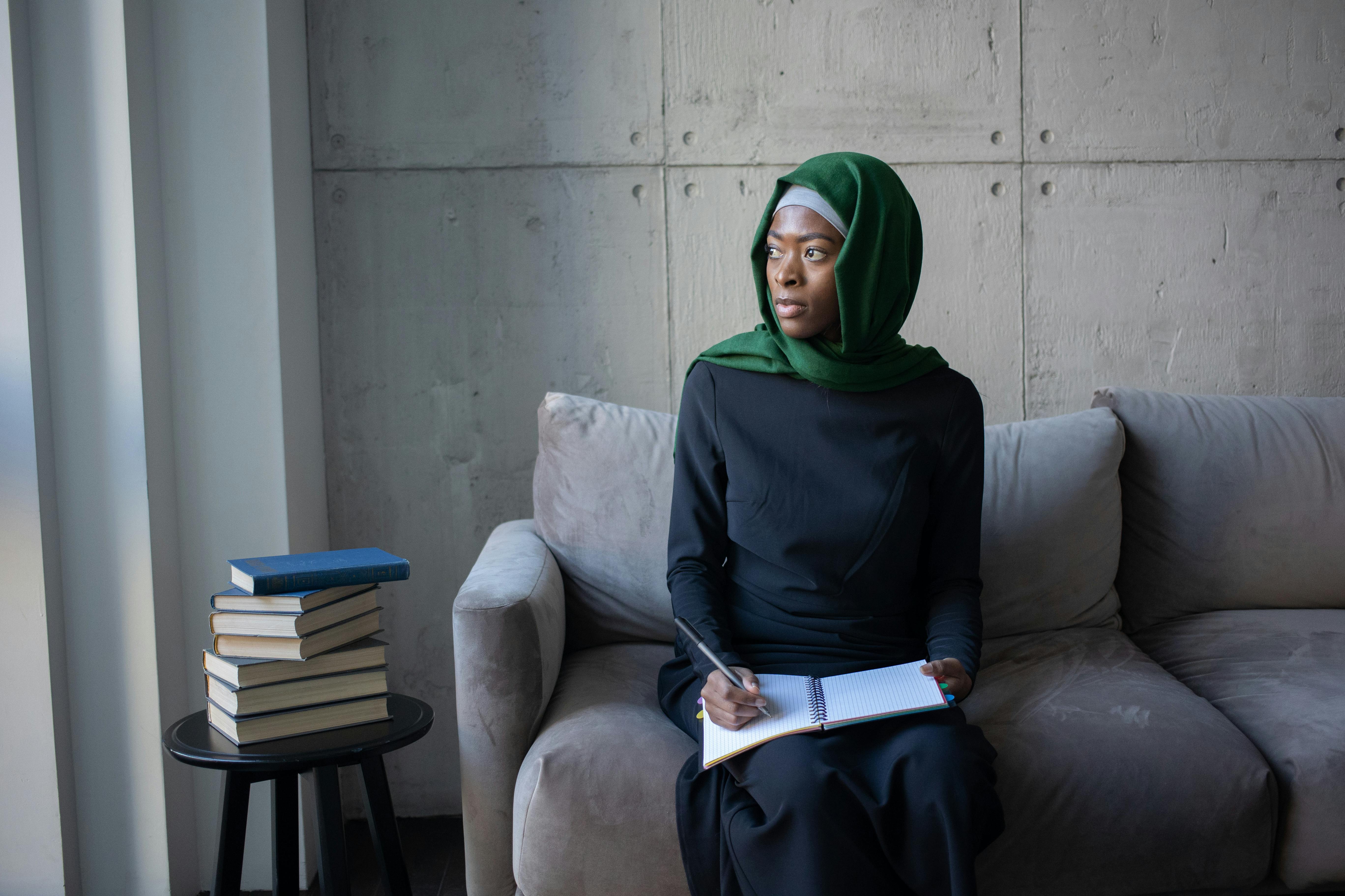 A female student in a hijab holding a pen and notebook on a grey sofa