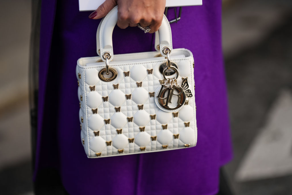 A woman in a purple dress holding a white and golden Dior Lady Dior bag.
